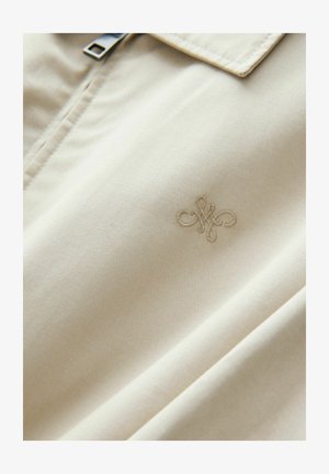 Svijetlosmeđi materijal s glatkom teksturom; sadrži suptilno izvezeni logo i srebrni patentni zatvarač na ovratniku.