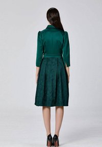 Femme se tenant tournée vers l'arrière, portant une robe mi-longue vert foncé à manches trois-quarts et des talons hauts noirs.