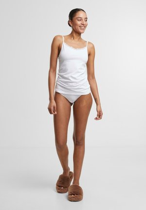 Weißes Baumwoll-Camisole mit dünnen Trägern und Spitzendetails, kombiniert mit weißen Slips und braunen, flauschigen Hausschuhen, das eine entspannte Passform zeigt.