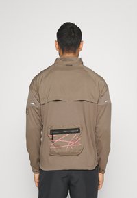 Bruine ritsjack met hoge kraag, gestructureerde stof en een achterzak met een roze lineair patroon en rits.