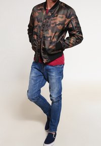 Kurtka bomber w kamuflażu w kolorze zielonym, brązowym i czarnym, z prążkowanym kołnierzem i mankietami, w połączeniu z niebieskimi dżinsami i granatowymi butami wsuwanymi.