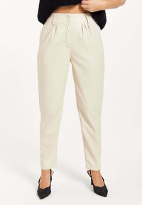 Cream pleated trousers with a high waist, two front pockets, and a smooth finish. Paired with black pointed-toe heels.