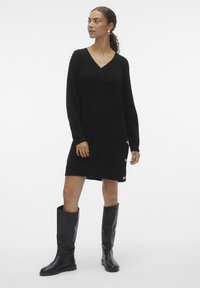 Robe en maille noire avec un décolleté en V, des manches longues et un détail de bouton sur le côté. Associée à des bottes en cuir noires jusqu'aux genoux.