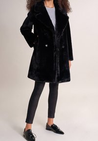 Černý kabát z umělé kožešiny s dvouřadým zapínáním, velkým klopovým límcem a dvěma předními kapsami, doplněný černými úzkými kalhotami a botami.