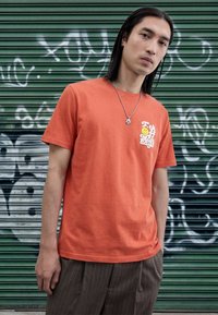 Orange bomulds t-shirt med rund halsudskæring, med et gult smiley-grafik og tekst på venstre brystområde. Bæreres med bukser med nålestriber.