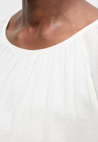 Κοντινό ενός ατόμου που φοράει λευκό, φαρδύ τοπ με στρογγυλή λαιμόκοψη και διακριτικές πιέτες.