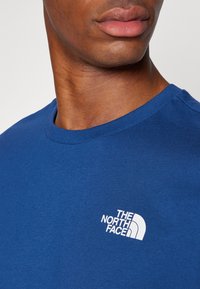 Zila kokvilnas T-krekls ar apaļu kakla izgriezumu. Uz kreisā krūšu sadaļas ir balta "The North Face" logo. Gluda tekstūra.