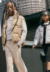 Twee kinderen lopen buiten in een beige puffervest, grijze trui en crèmekleurige broek; het tweede kind draagt een zilveren pufferjack en een zwarte rok.