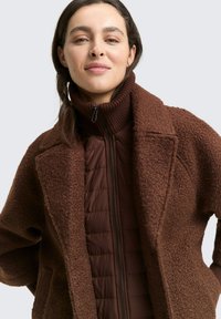 Bruine fleece buitenjas met een gestructureerd oppervlak, gelegen over een bruine gewatteerde binnenjas met een hoge kraag en ritsluiting.
