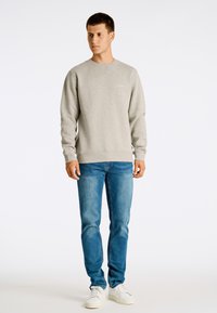 Felpa grigia con collo a rondine e polsini a costine, abbinata a jeans blu e sneakers bianche. Tessuto morbido con dettagli di branding minimi.