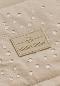 Beige szövet vízcseppekkel és egy texturált gumicimkével, amelyen a "JOHNNY URBAN" egyszerű betűtípussal van gravírozva.