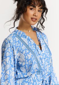 Robe florale bleue avec des motifs botaniques blancs, décolleté volanté et détails sur le buste, avec une coupe décontractée et une taille à cordon.