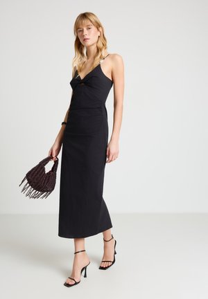 Donna con un vestito midi nero senza maniche con spalline sottili, che tiene una borsa scura con perline, indossa sandali neri con tacco alto, in piedi su uno sfondo bianco.