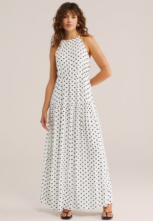 Kobieta ubrana w białą sukienkę maxi bez rękawów w czarne kropki, stoi z jedną ręką na biodrze, nosi czarne sandały.