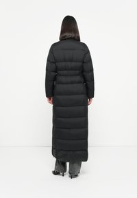 Dlhý čierny puff coat s priliehajúcim pásom a horizontálne prešívanými sekciami, siahajúci po pol lýtok, vyrobený z hladkého, ľahkého materiálu.