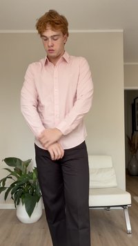 Camisa de mangas longas cor-de-rosa clara com botões, combinada com calças pretas. Apresenta um corte descontraído e textura suave. Ambiente interior com planta verde.