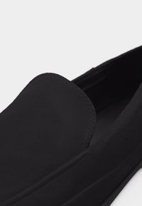 Κοντινή λήψη ενός μαύρου παπουτσιού slip-on με στρογγυλεμένη μύτη και απαλό, ματ φινίρισμα σε λευκό φόντο.