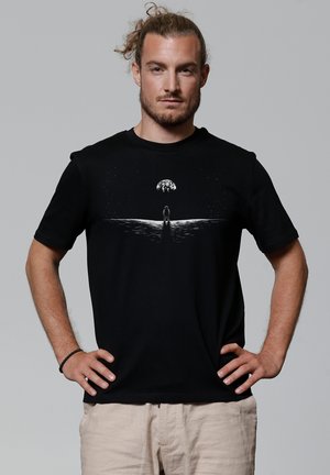 Zwarte katoen T-shirt met korte mouwen met een grafisch ontwerp van een figuur en een hemelse lichaam, geplaatst tegen een getextureerde achtergrond.