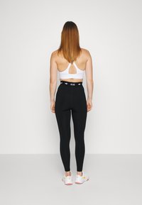 Black high-waisted leggings with a branded waistband, paired with a white sports bra featuring a back cutout design; worn with white athletic shoes.