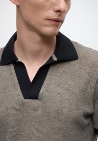 Adīts polo krekls ar melnu apkakli, teksturētu audumu bēšā krāsā ar ģeometrisku rakstu un pusvērstu pogu aizdari kakla daļā.