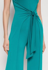 Mono en tonos verdes con tejido ligero y un detalle de lazo en la cintura, que presenta una silueta fluida y piernas de pantalón alargadas.