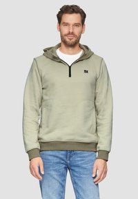 Lichtgroene zip-up hoodie met een voorvak, ribgebreide boorden en onderrand, met een logo op de borst en een capuchon. Draagt met blauwe jeans.