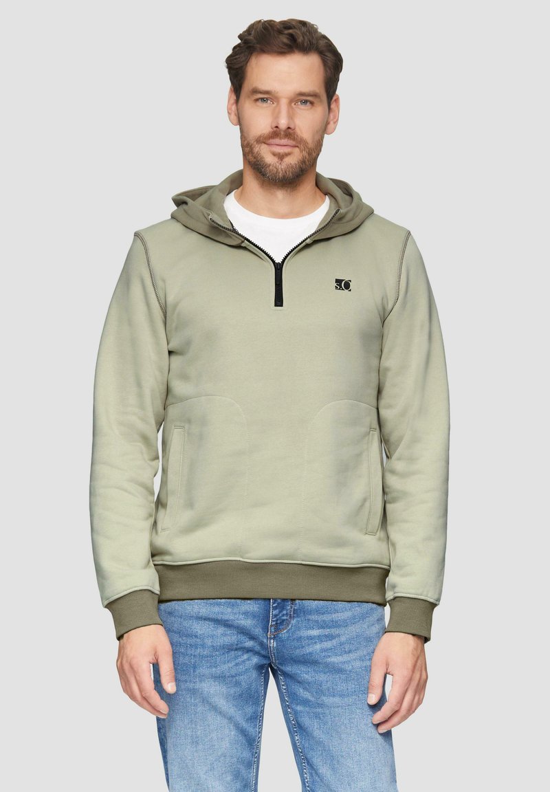 Lichtgroene zip-up hoodie met een voorvak, ribgebreide boorden en onderrand, met een logo op de borst en een capuchon. Draagt met blauwe jeans.