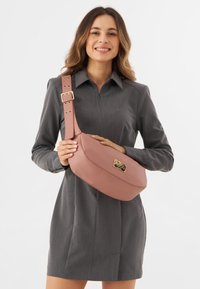 Borsa a tracolla in finta pelle rosa con forma arrotondata, tracolla regolabile e dettagli in oro. Texture liscia con particolare logo.