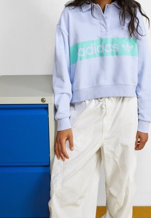 Osoba nosząca jasnofioletową bluzę Adidas i luźne białe spodnie, stojąca obok niebiesko-beżowej szafy na dokumenty.