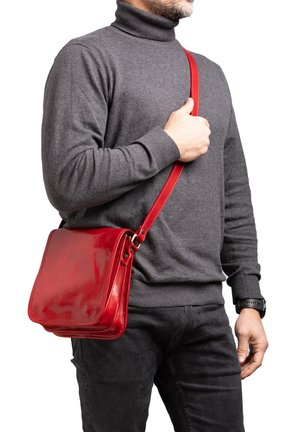 Man in grijze coltrui die de riem vasthoudt van een glanzende rode leren schoudertas tegen een witte achtergrond.