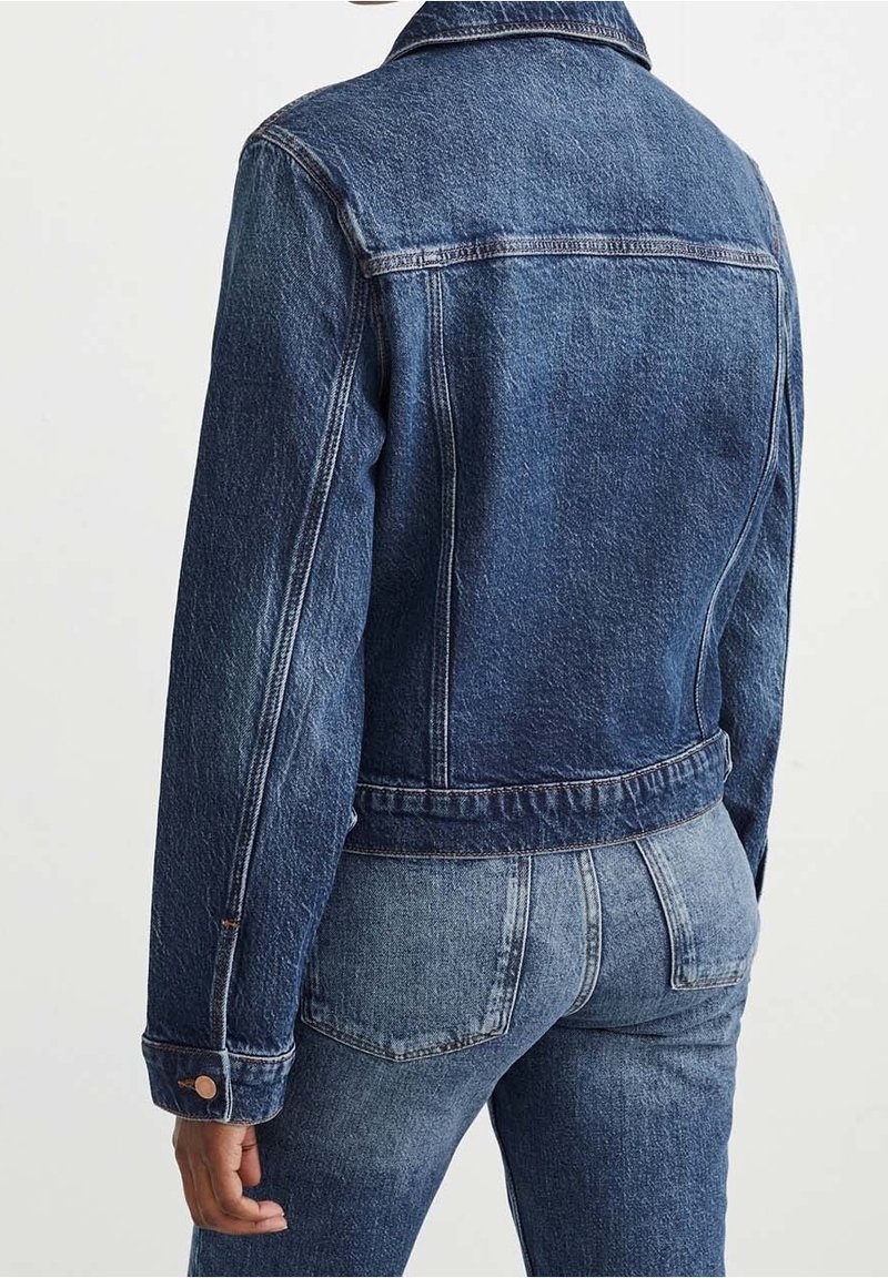 Personne portant une veste en jean bleu foncé et un jean bleu, vue de dos sur un fond clair uni.