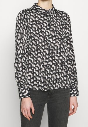 Woman wearing black and white abstract patterned long-sleeve button-up shirt with black jeans against a plain background.