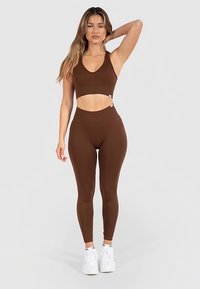 Brassière de sport marron et leggings taille haute, design ajusté, tissu texturé, assortis à des baskets blanches, détail de marque discret.