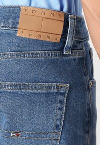 Niebieskie dżinsy z denimu z beżową skórzaną łatą oznaczoną "TOMMY JEANS". Charakteryzują się wyraźnymi szwami oraz małym haftowanym logo na tylnej kieszeni.