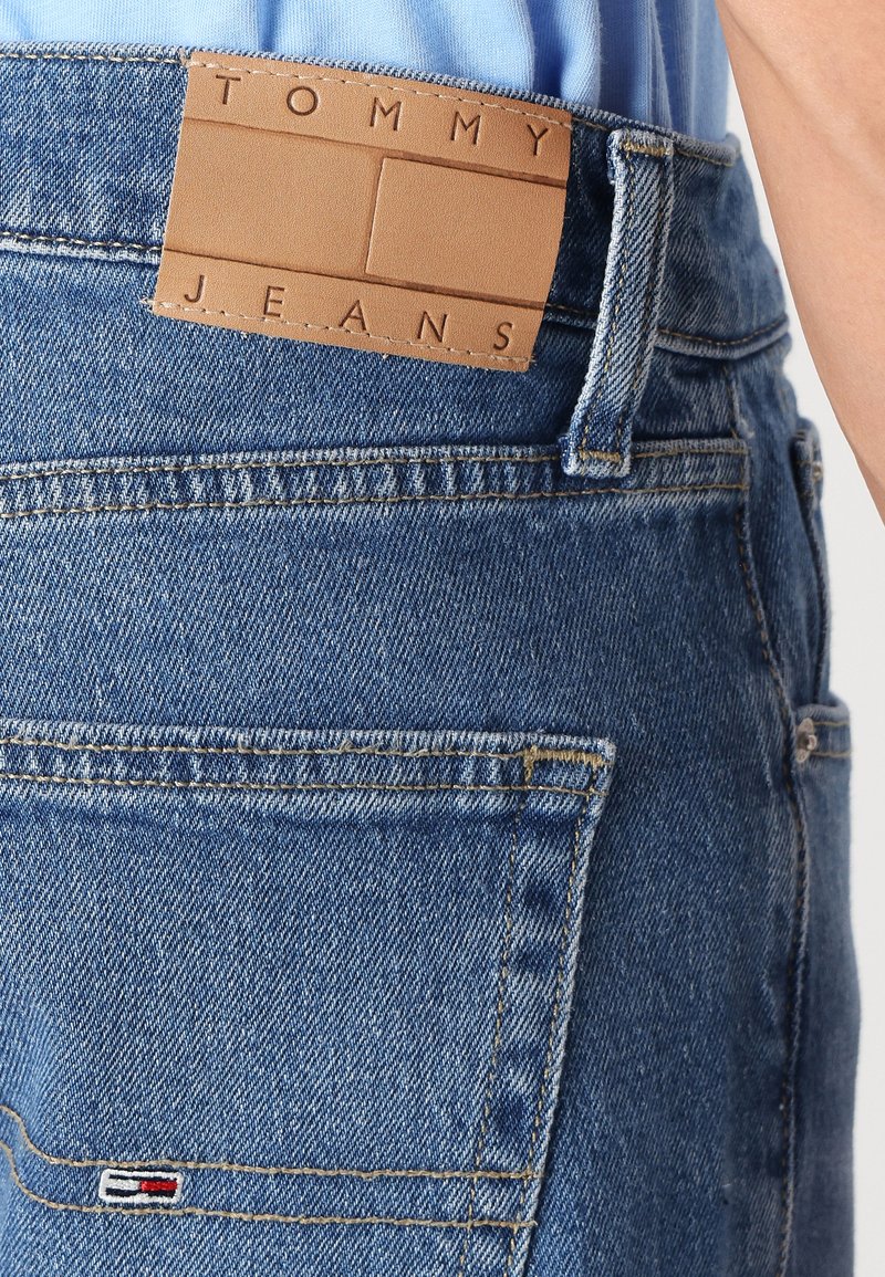 Blaue Jeans aus Denim mit einem hellbraunen Lederpatch, auf dem "TOMMY JEANS" steht. Mit auffälligen Nähten und einem kleinen gestickten Logo auf der Gesäßtasche.