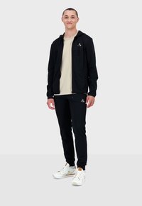 Giacca sportiva nera con chiusura a zip, abbinata a pantaloni joggers neri. Il modello indossa una maglietta beige e sneaker bianche con dettagli dorati.
