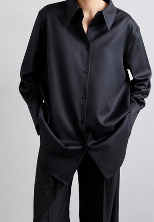 Persoon die een oversized zwart overhemd met wijde kraag draagt en bijpassende zwarte broek, handen in de zakken tegen een effen achtergrond.
