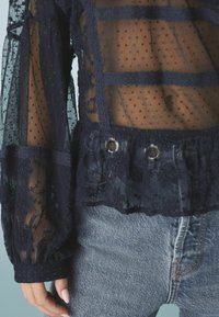 Blouse en voile marine avec un motif à pois et des accents en dentelle florale. Présente des volants superposés et des détails en métal argenté à la taille. Portée avec un denim clair.
