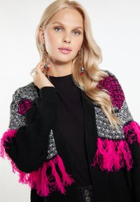 Svart kappa med rosa fransar och texturerade ärmar. Har en blandning av material, med en enfärgad svart kropp och mönstrade detaljer.