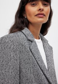 Houndstooth-kuvioinen bleiseri mustavalkoisena, jossa on leikattu kaulus. Kangas näyttää teksturoidulta ja siinä on klassinen, räätälöity istuvuus.