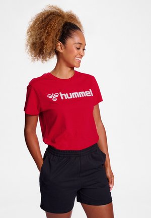 Усмихната жена с къдрава коса, облечена с червена тениска Hummel и черни къси панталони, на изчистен бял фон.