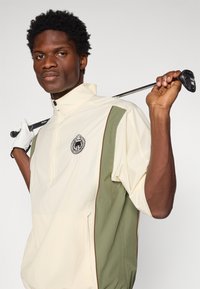 Golf jaka smilškrāsā un olīvzaļā, ar daļēju rāvējslēdzēja dizainu, sānu kabatām un logo akcentu uz krūtīm. Strukturēta auduma apdare.