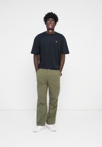 Homme avec les mains dans les poches portant un t-shirt noir avec un logo jaune, un pantalon cargo olive et des baskets blanches, se tenant devant un fond blanc uni.