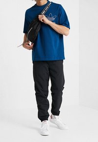 Blå bomullströja med fronttryck, svart nylonmidjeväska, svarta byxor och vita sneakers med röda detaljer. Avslappnad, ledig passform.