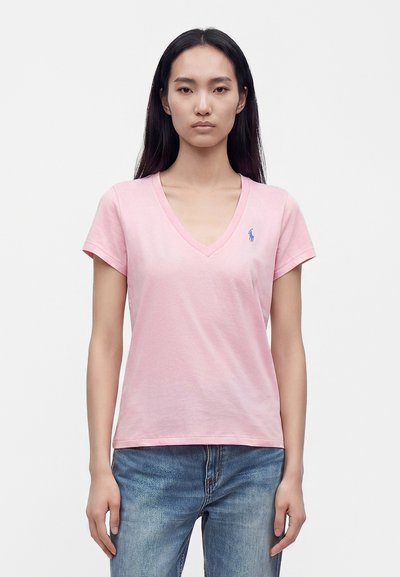 Γυναίκα που φοράει ανοιχτό ροζ T-shirt με V λαιμόκοψη, με μικρό μπλε λογότυπο στο αριστερό στήθος και μπλε τζιν, στέκεται μπροστά σε απλό φόντο.
