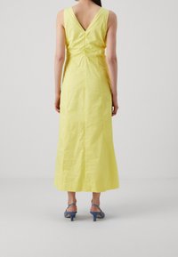 Żółta bawełniana sukienka maxi z dekoltem w kształcie V, marszczeniem w talii i elegancką sylwetką. Zestawiona z niebieskimi sandałami na obcasie.