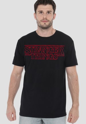 Man wearing a black T-shirt with red "Stranger Things" text, standing against a plain light gray background.