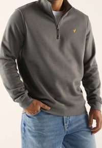 Sudadera gris con cremallera y textura suave, con cuello alto y un pequeño logo amarillo en el pecho. Combinada con vaqueros azules.