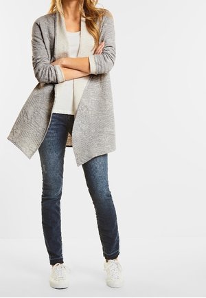 Cárdigan gris claro con frente abierto, mangas largas y tejido texturizado, combinado con una camisa blanca, jeans de mezclilla oscuros y zapatillas blancas.