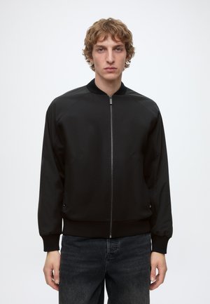 Jeune homme aux cheveux bouclés portant une veste noire zippée et un jean foncé, debout devant un fond blanc uni.